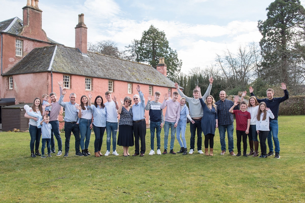 Family of twenty-one, from a baby to retired grandparents, waving at the camera on a lawn in front of a peach coloured house