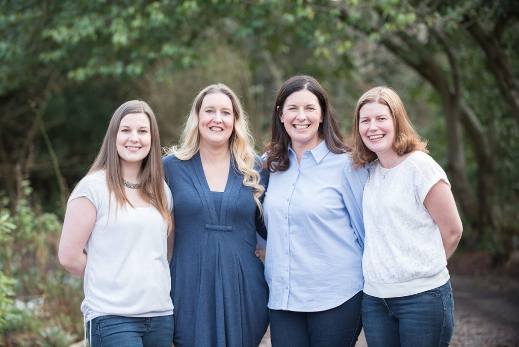 Four women smiling with their arms around each other, dressed in blue and white, in a family portrait photograph in woodland