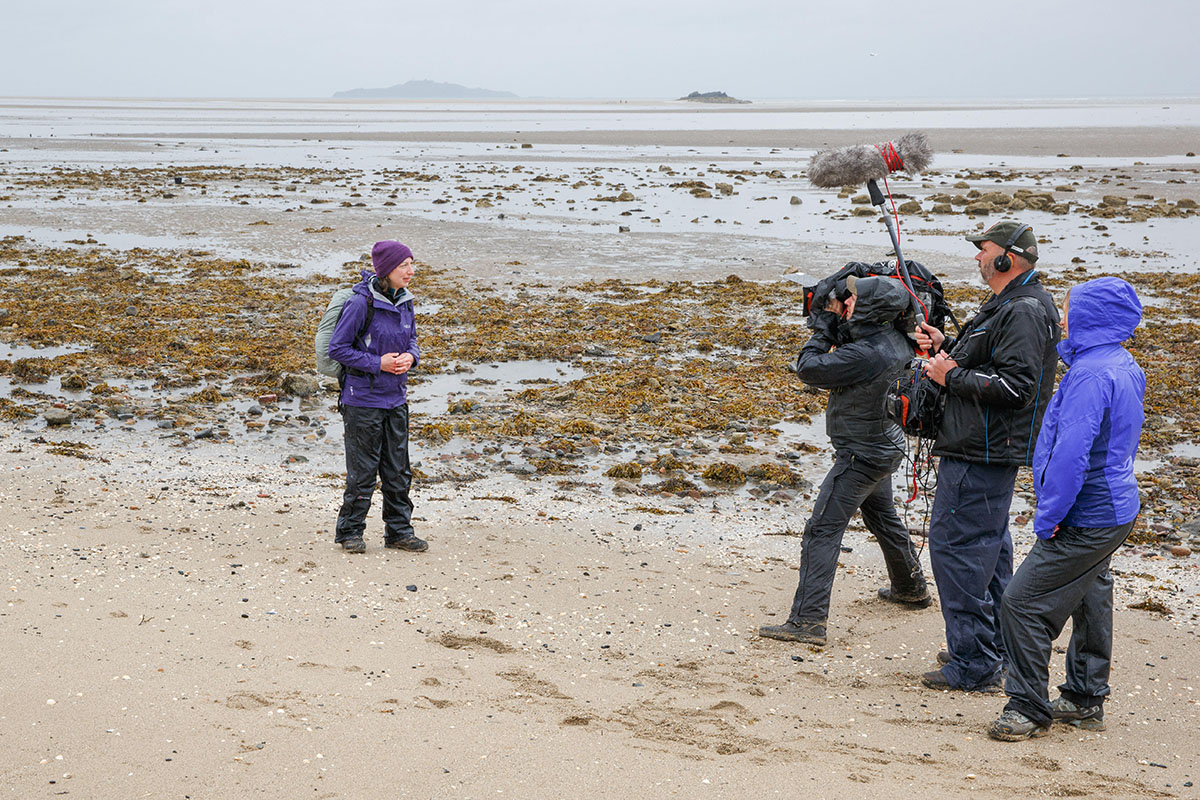 Guest blogger - woman dressed in wet weather clothing on a beach with sand and sea weed with a three person film crew