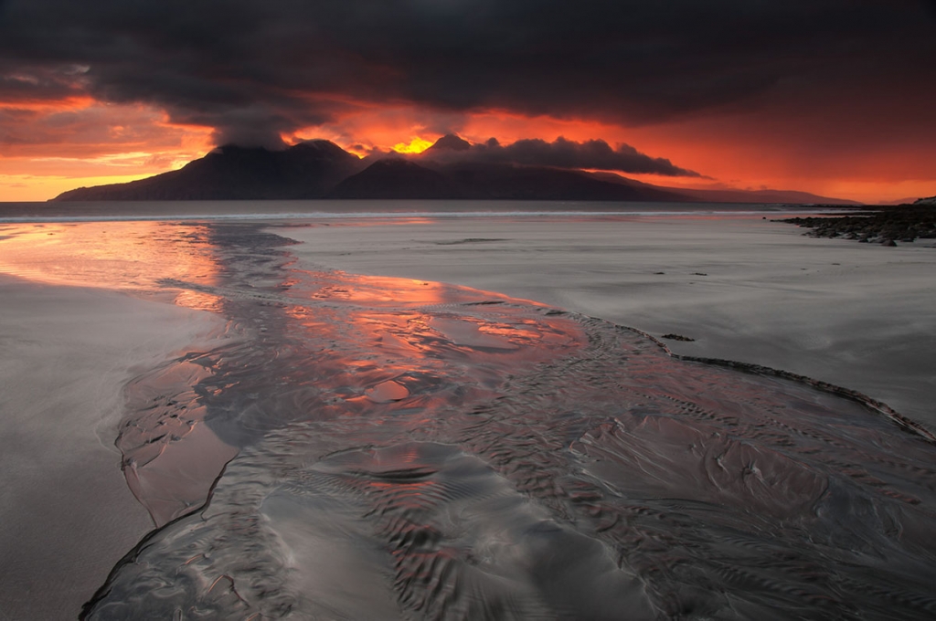 Guest speaker - dramatic orange and yellow sunset behind a mountainous island with the sea and a beach in the foreground
