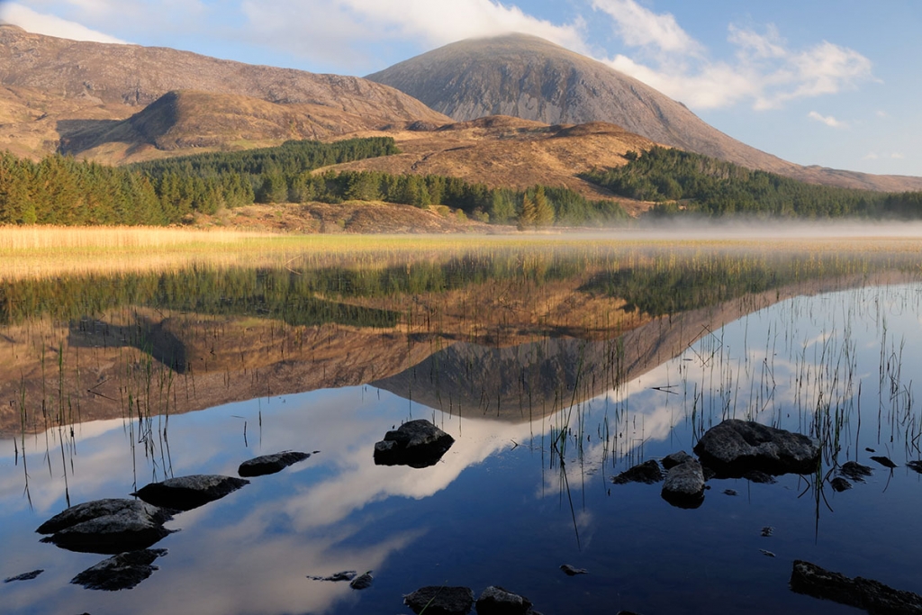 Isle of Skye photography - mountain, hills and forest under blue sky, reflected in a lake with rocks in the foreground