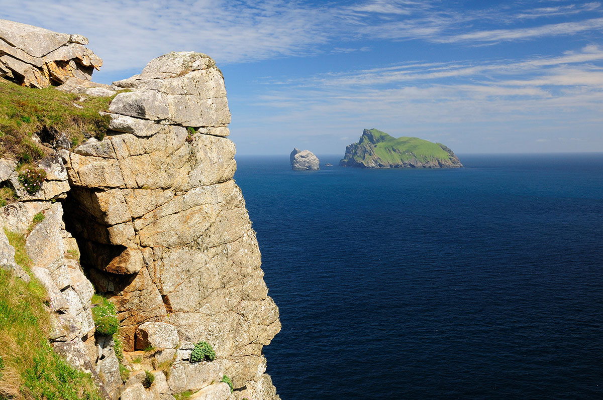 Distant green Scottish island and sea stacks in a blue sea under a blue sky, with a cliff face in the foreground