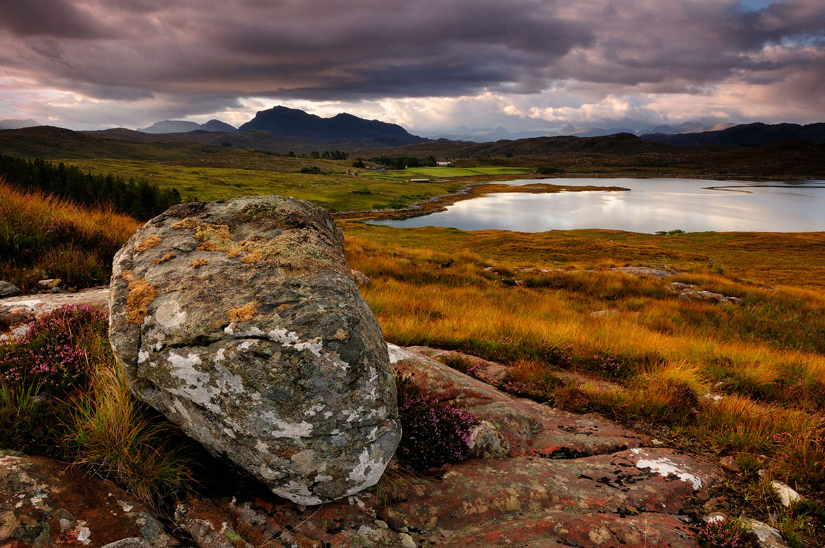 Large grey boulder on top of granite on a grass slope overlooking a bay with mountains beyond, under a dramatic cloudy sky