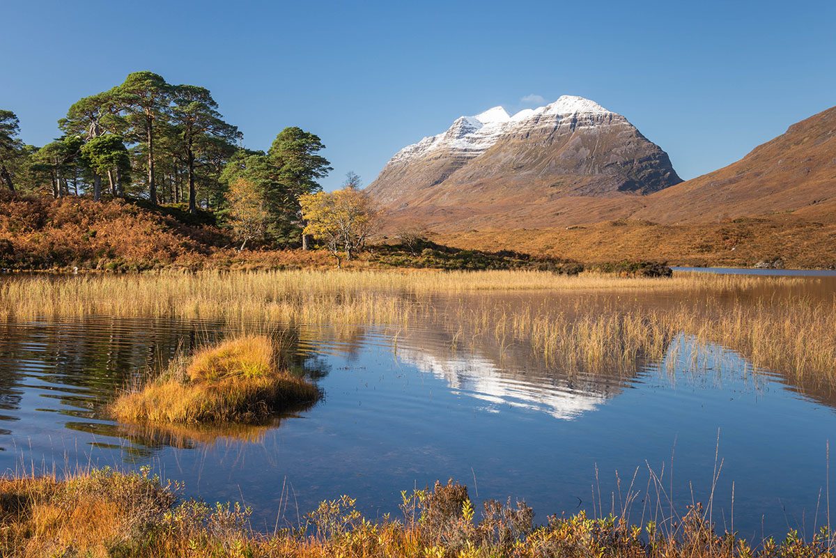 Steep sided mountain with snow on its summit behind a Scottish loch and native woodland in autumn colours, beneath blue sky