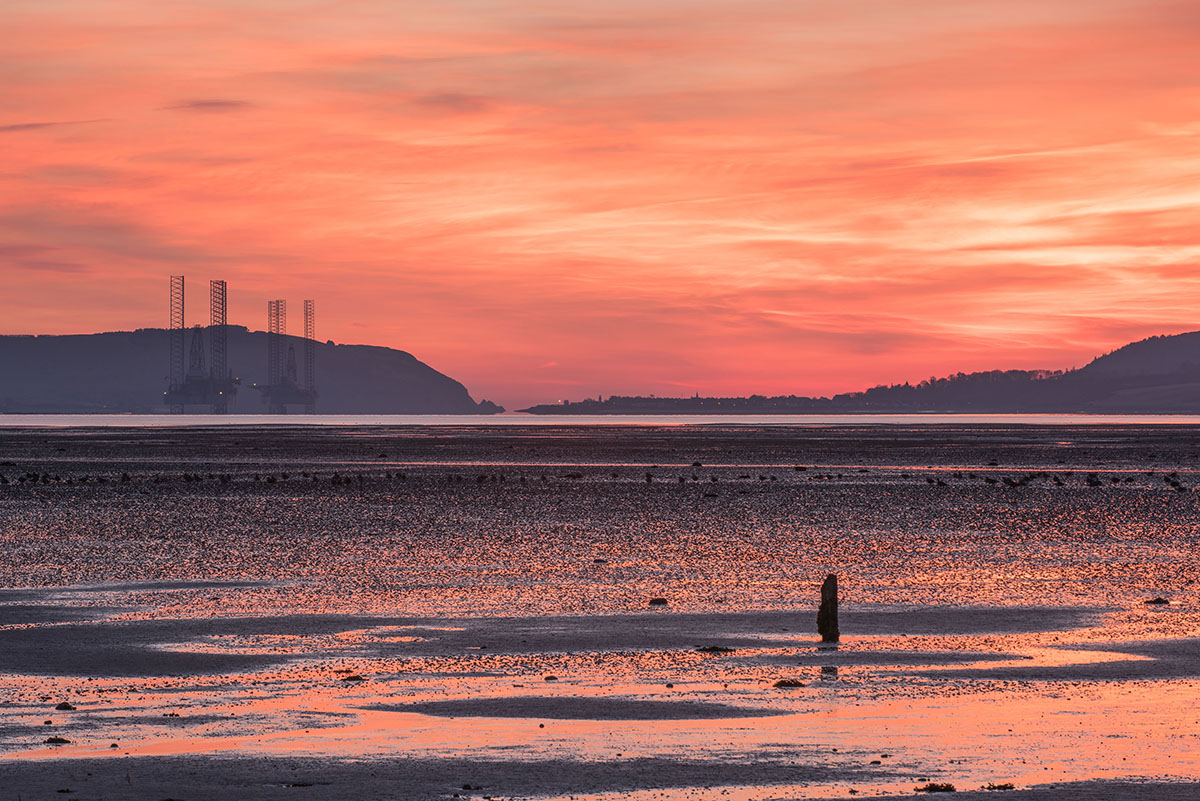 Black Isle photography - post sticking out of mudflat at low tide with oil rigs and hills in the distance under an orange sky
