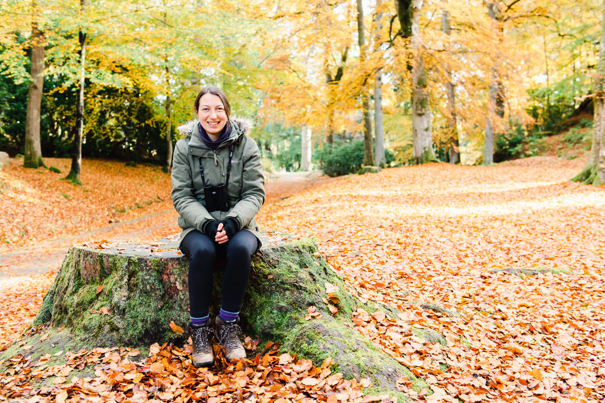Female wearing a green jacket and black leggings, smiling and sitting on a large tree stump in a mature woodland in autumn