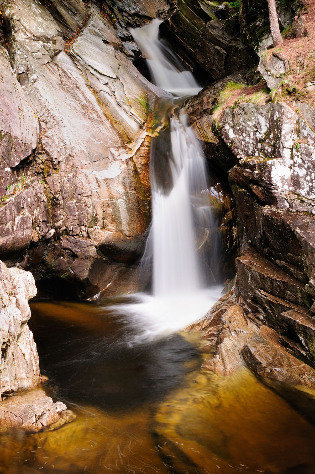 Waterfall photographed in slow motion, in a small gorge with smooth grey rocks on either side and dark pool at the base