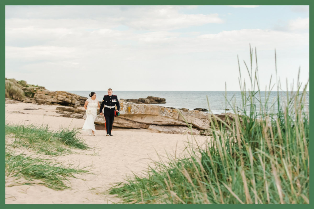 Wedding photograph of a bride and groom walking hand-in-hand on a sandy beach with rocks and marram grass