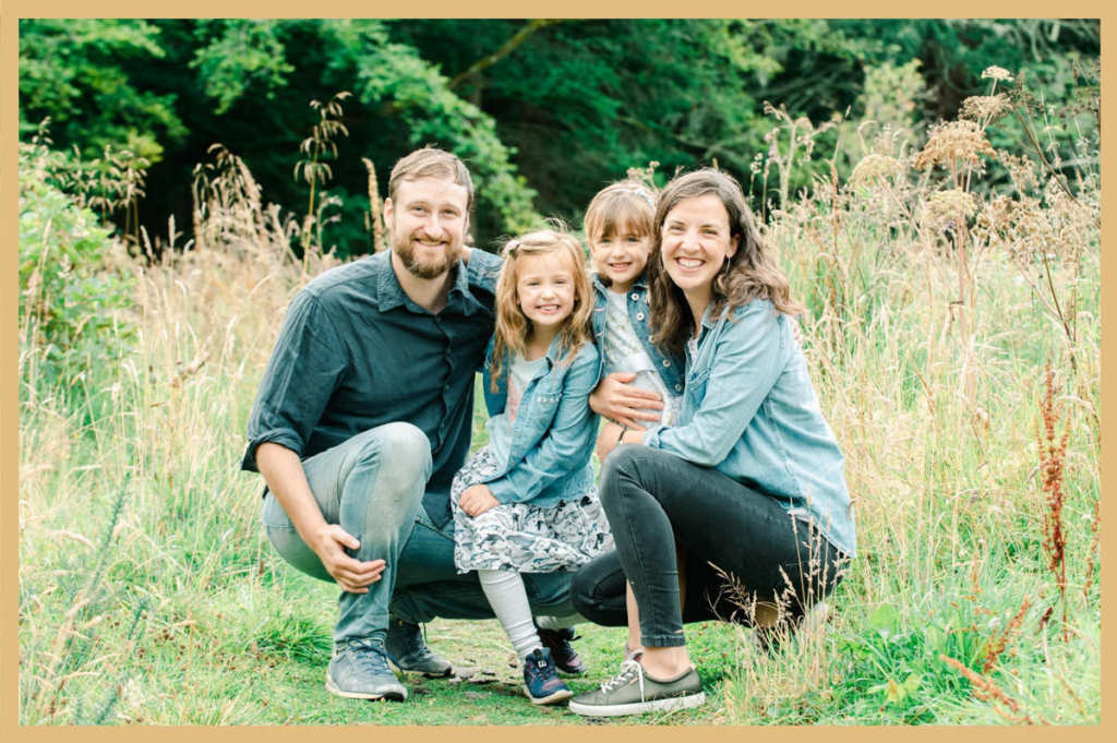 Parents crouching beside their two young daughters for a family photograph in tall grass in front of an Inverness woodland