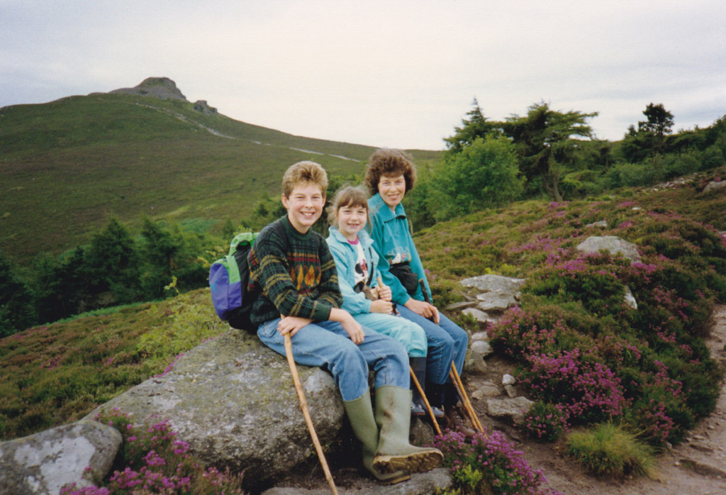 A family photo of a mum, a boy and a girl sitting on a rock by the side of a footpath with trees and a hill summit beyond
