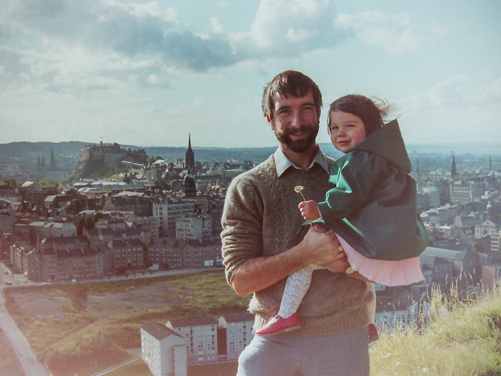 A photo of a young dad holding his toddler age daughter with a city skyline with a castle and spire behind them