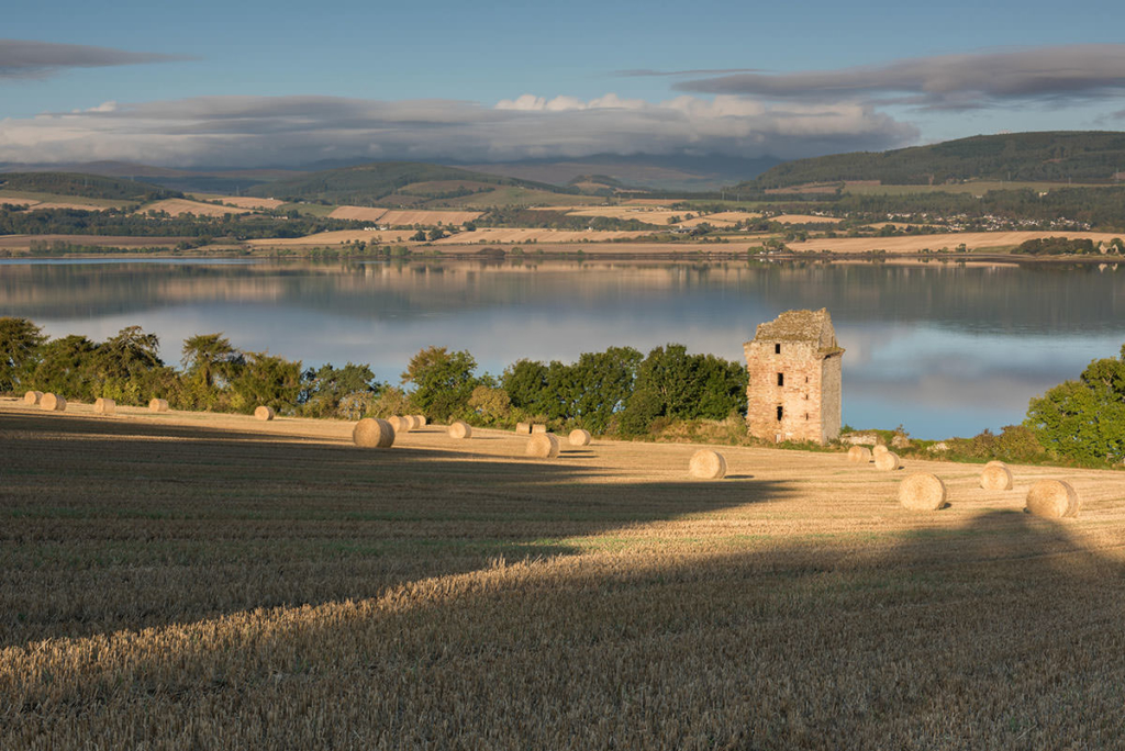 Ruined Scottish Highlands castle in field with straw bales and long shadows in foreground, with a lake and hills beyond
