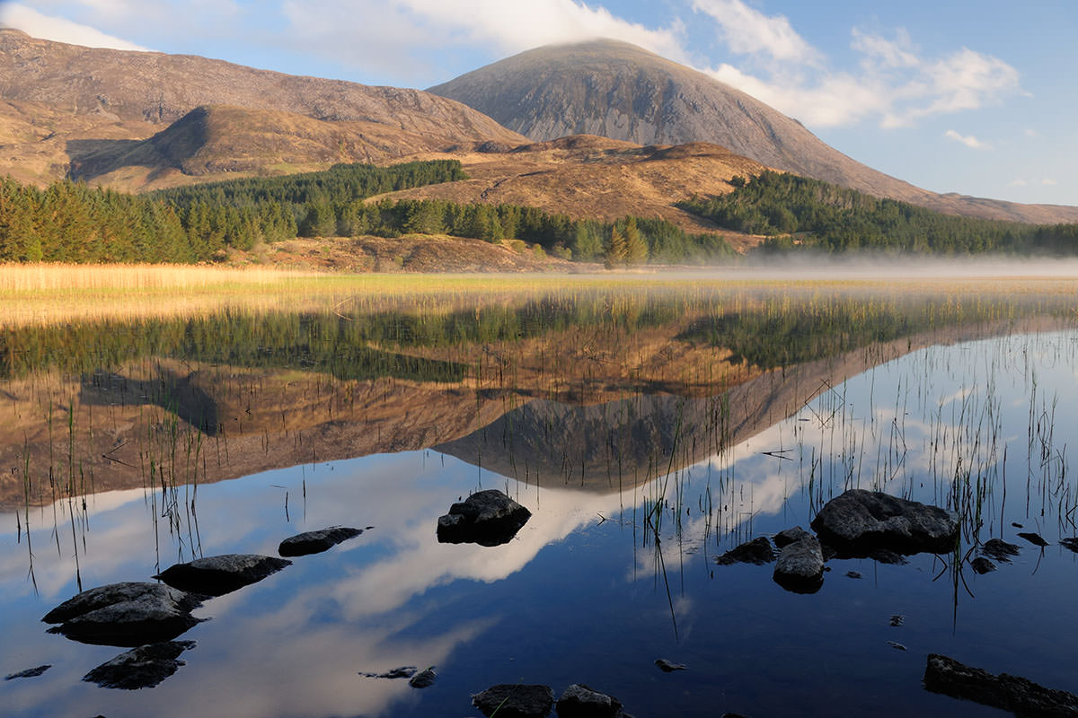 A Scottish Highland landscape with a mountain, hills and forest reflected in a lake with rocks in the foreground