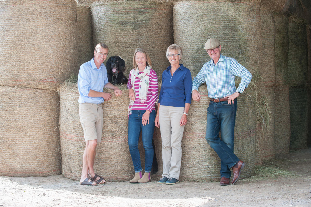 Adult family in outdoor clothing with a black dog standing in front of stacked straw bales