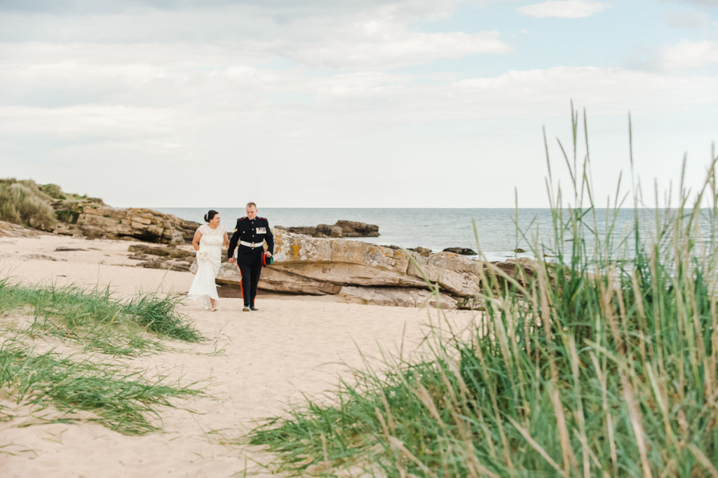 Wedding photograph of a bride and groom walking hand-in-hand on a sandy beach, beside large rocks and marram grass