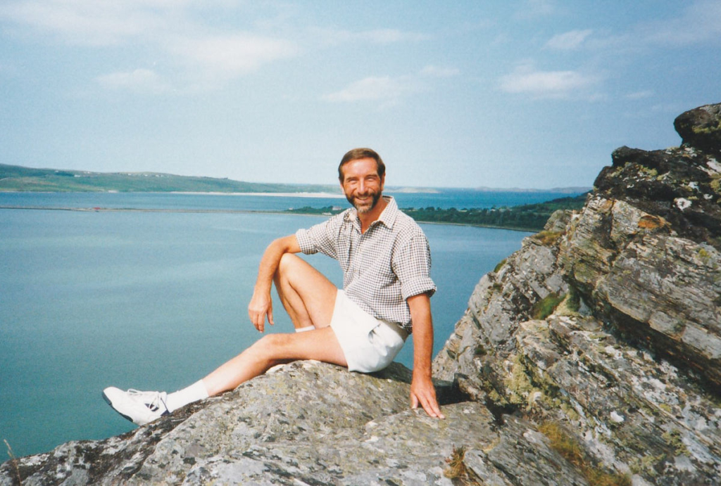 Photograph of a man wearing shorts sitting on a rock and smiling, with the sea and a causeway in the distance