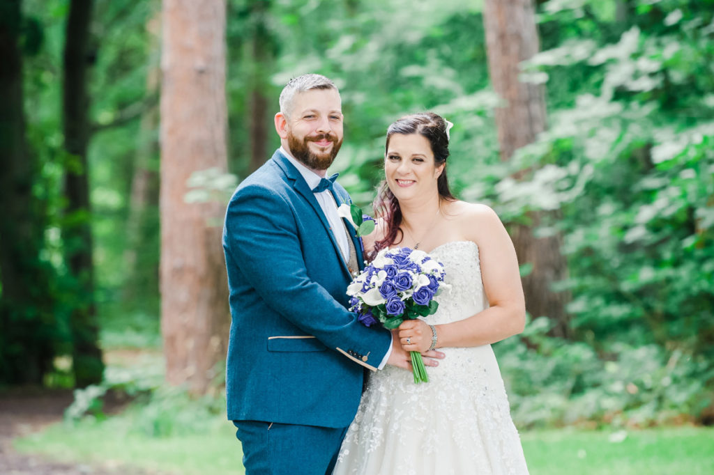 A groom in a blue suit and a bride in a white dress smiling and standing in front of tall tree trunks and green vegetation