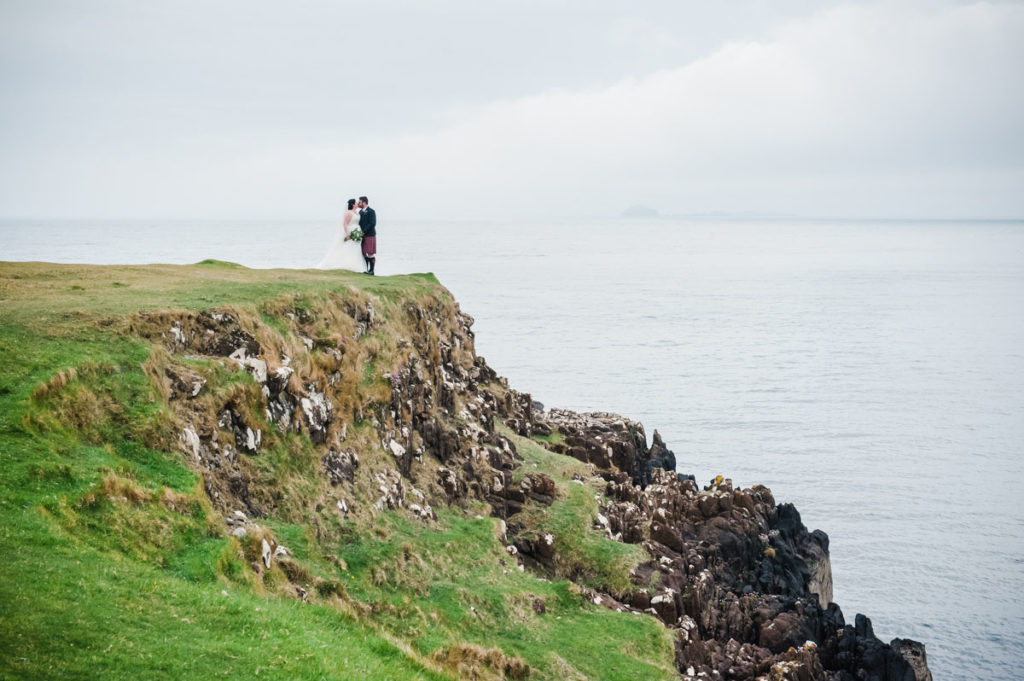 A bride in a white dress and a groom in a red kilt standing on top of a green cliff above the sea with clouds in the sky
