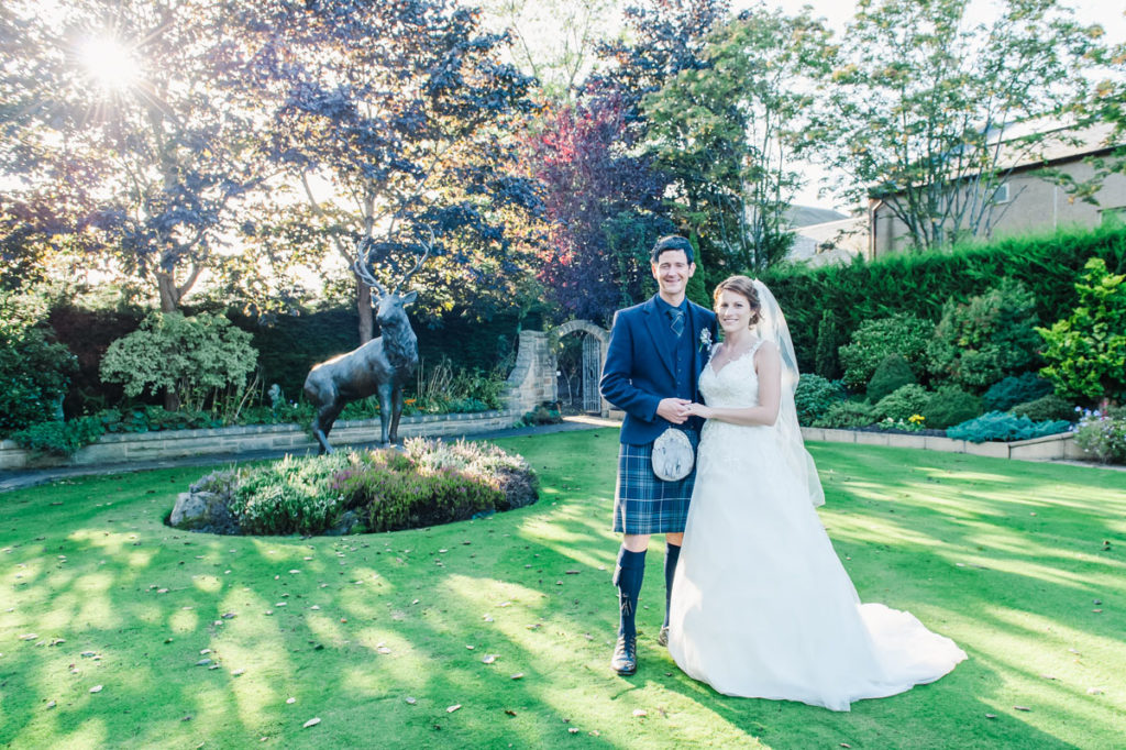 A bride and groom holding hands and standing on a lawn next to a statue of a stag with sunlight coming through the trees