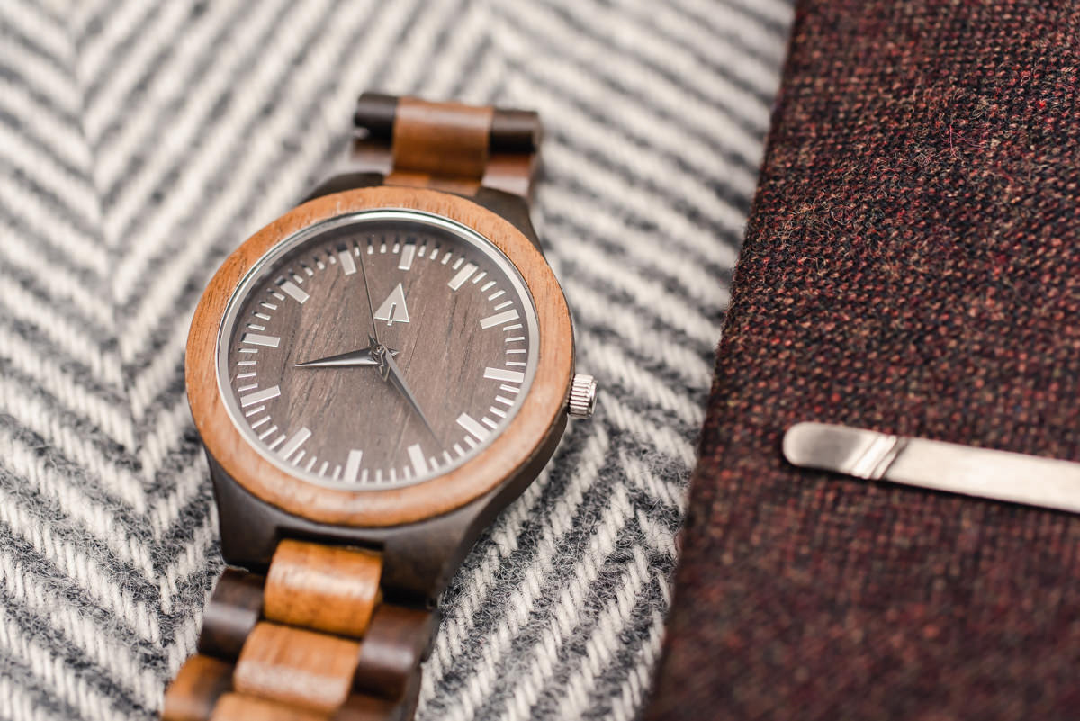 A brown watch next to a brown tie with a metal tie clip on top of a hatched brown and white background
