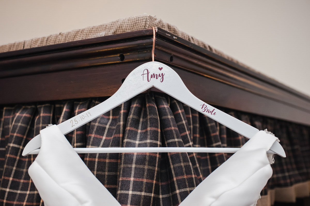 A coat hanger with 'Amy' on it, hanging on a wooden four poster bed with dark curtains, with the top of a white dress showing