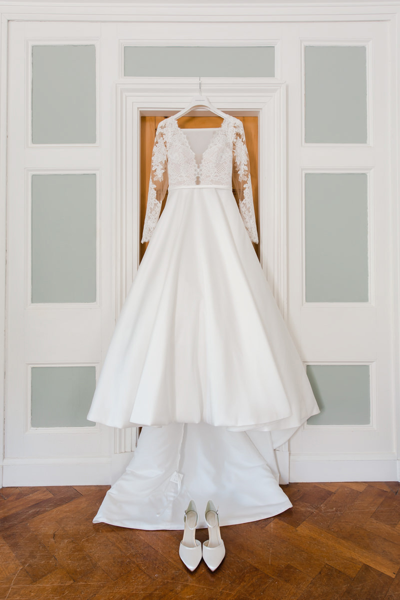 An elegant white wedding dress and shoes hanging in a doorway in a room with a wooden floor and white and green panelling