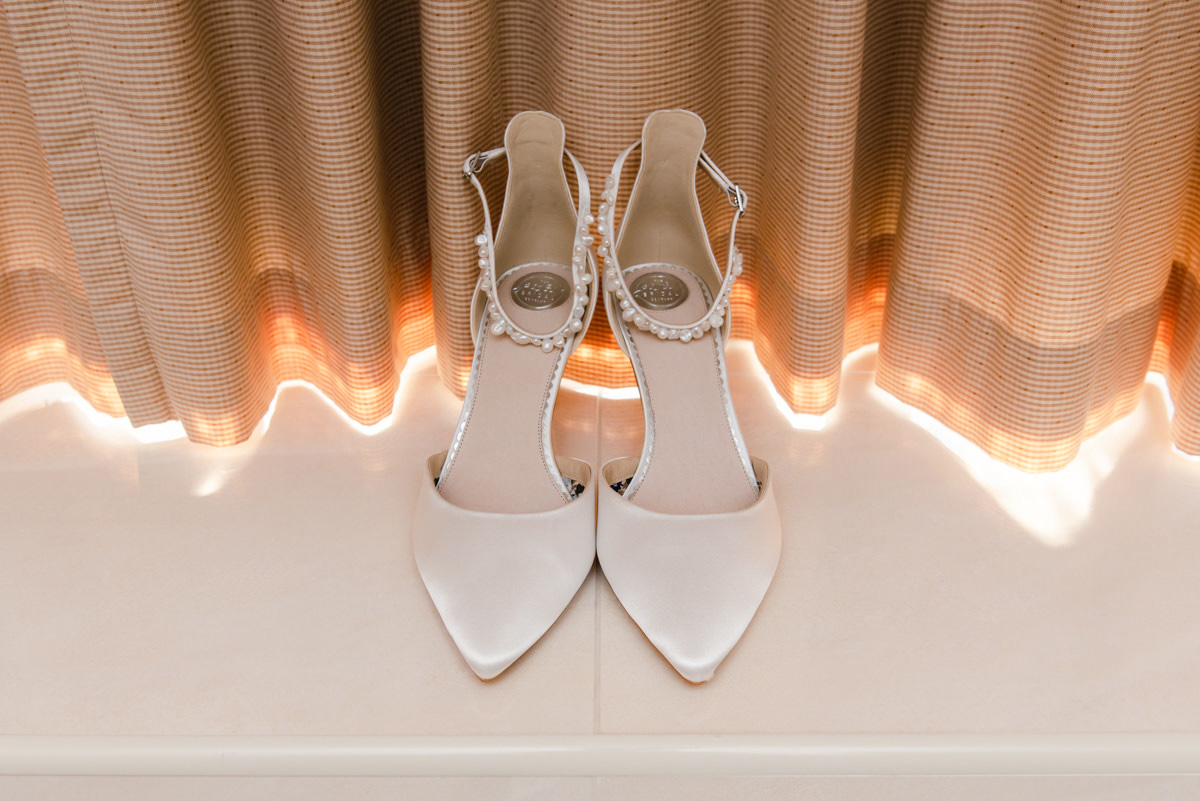 A pair of white bridal shoes on a tiled surface in front of cream curtains