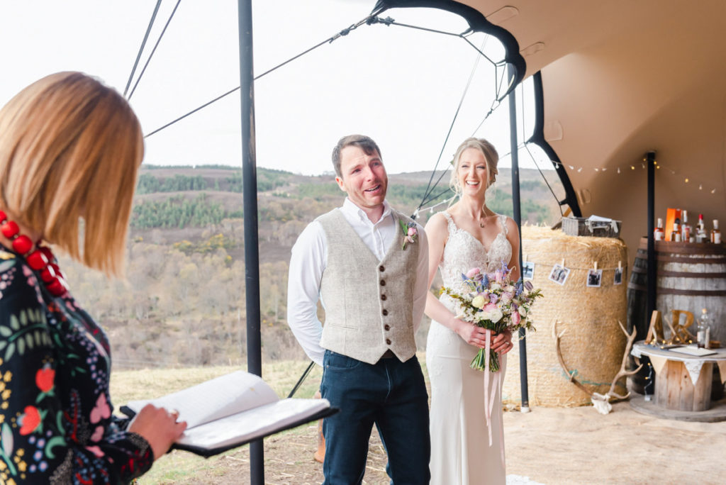 A groom looking tearful next to his laughing bride beside a wedding celebrant in a stretch tent with hills beyond