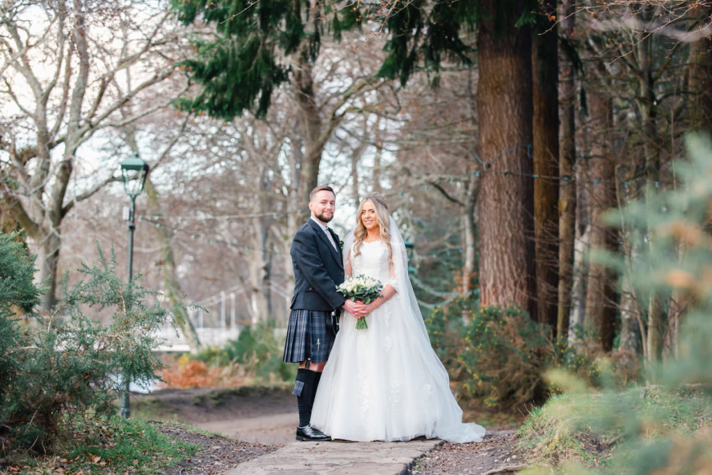 A Caucasian bride and groom standing together on a woodland path in front of a traditional lamppost, bushes and tall trees
