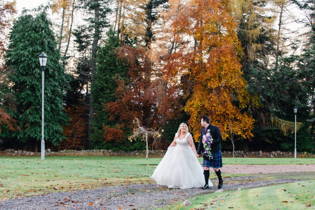 A bride in a white dress laughing and holding hands with a groom in a kilt walking on a path in front of lampposts and trees