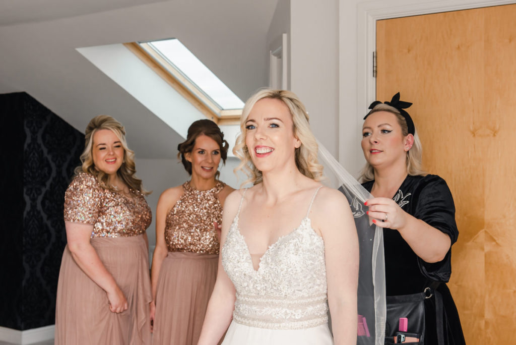 A blonde bride in a white wedding dress having her veil fitted by a woman in black, in front of two bridesmaids in pink