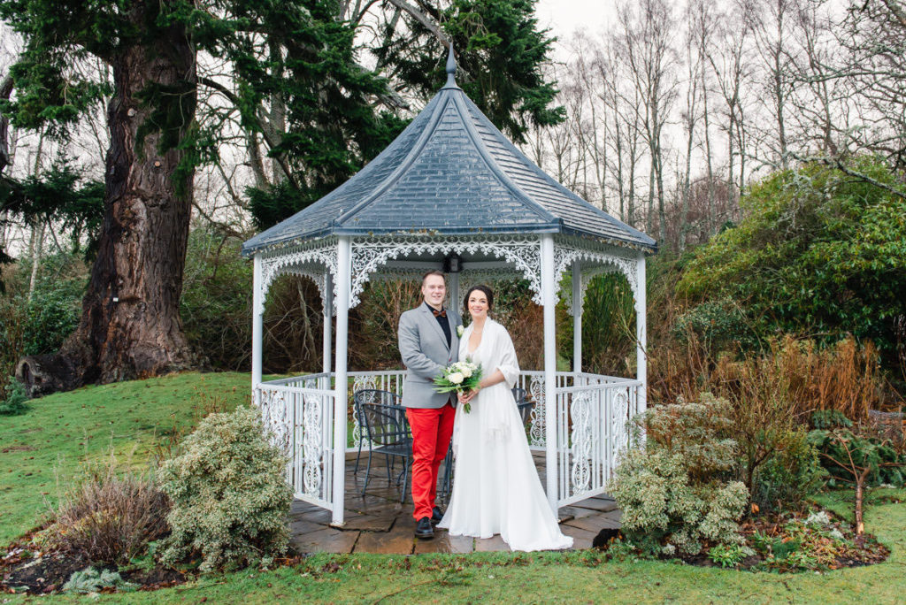 A bride in a white dress and a groom in a grey suit jacket and red trousers in a pergola in a garden with plants and trees