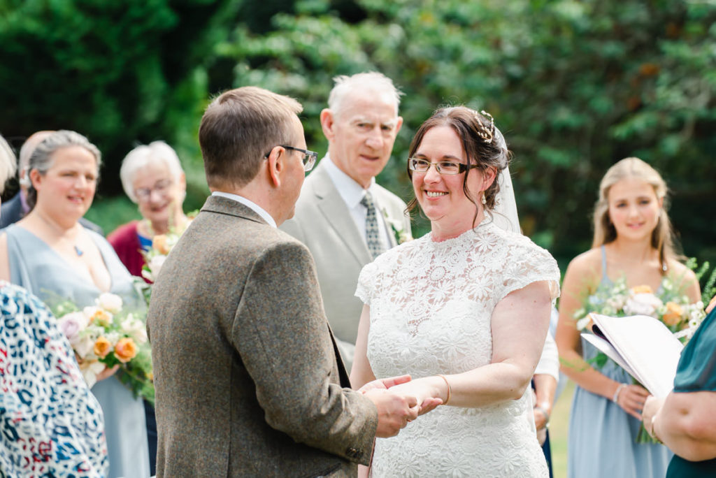 Caucasian middle aged bride and groom exchanging rings in an outdoor wedding ceremony with guests and trees in the background