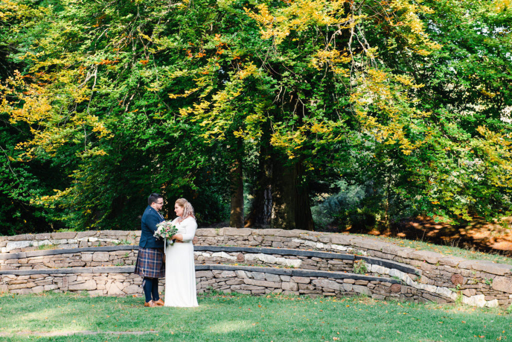 A bride and groom in a dress and kilt standing facing each other on grass in front of a curved stone wall with trees behind