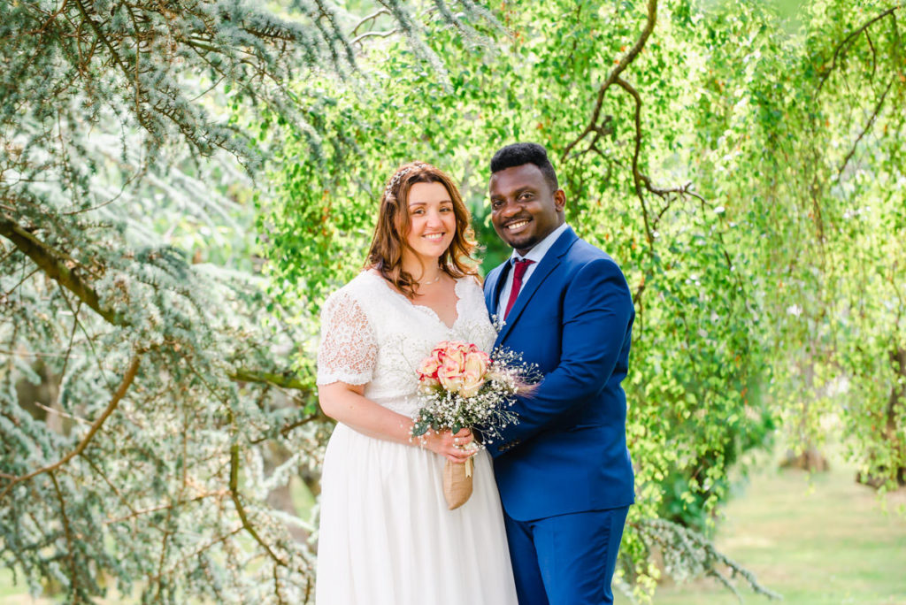 A Caucasian bride in a white dress holding a flowers and standing beside a man of colour in a blue suit, in front of trees