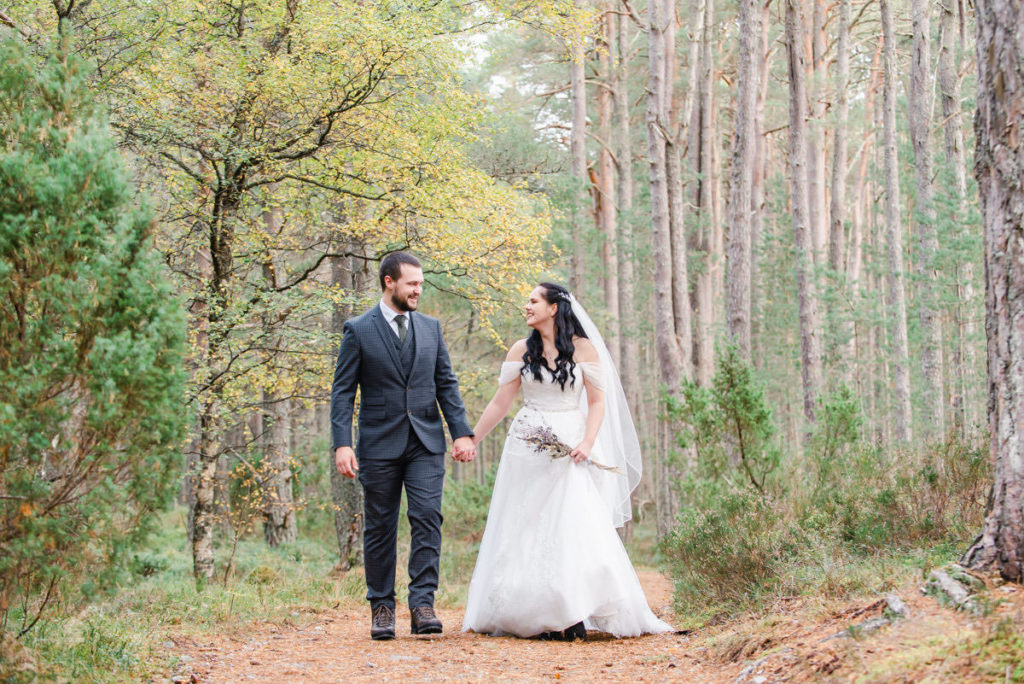 A Caucasian bride and groom holding hands and smiling at each other walking on a woodland path next to trees