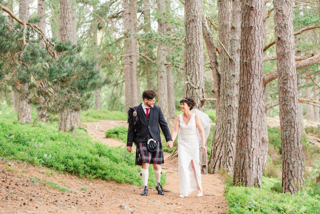 A Caucasian bride and groom in a white dress and kilt holding hands and walking on a woodland path in front of tall trees