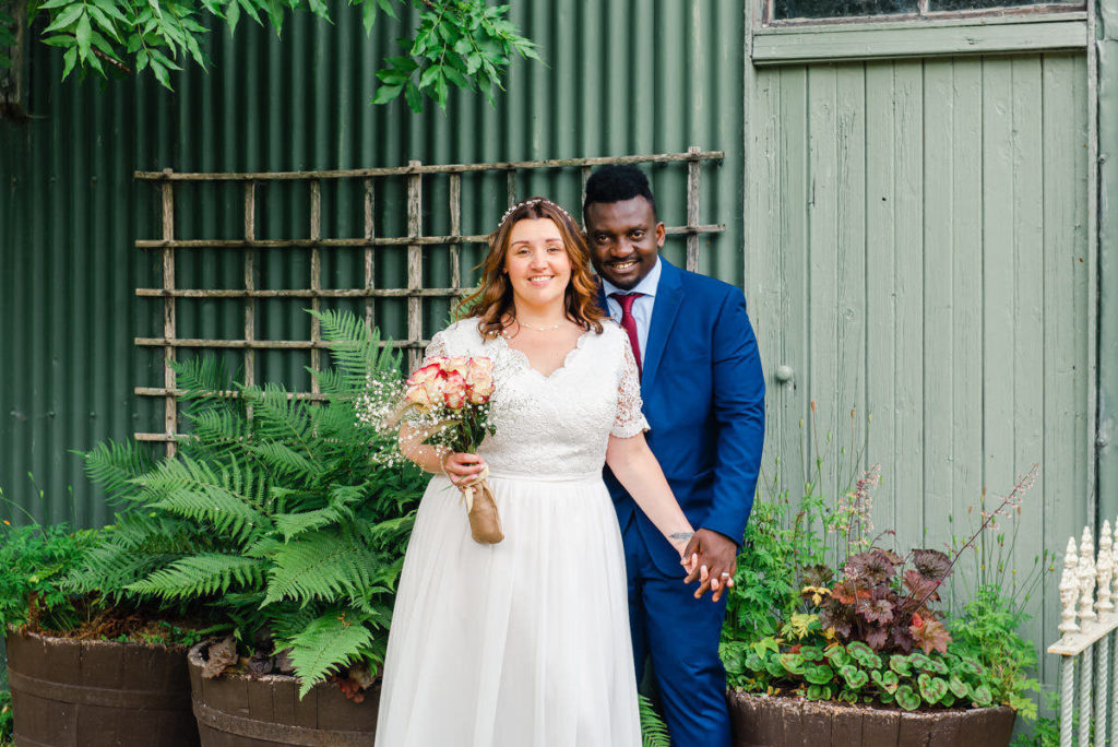 A Caucasian bride in a white dress holding hands with a groom of African origin, standing in front of a green shed and shrubs