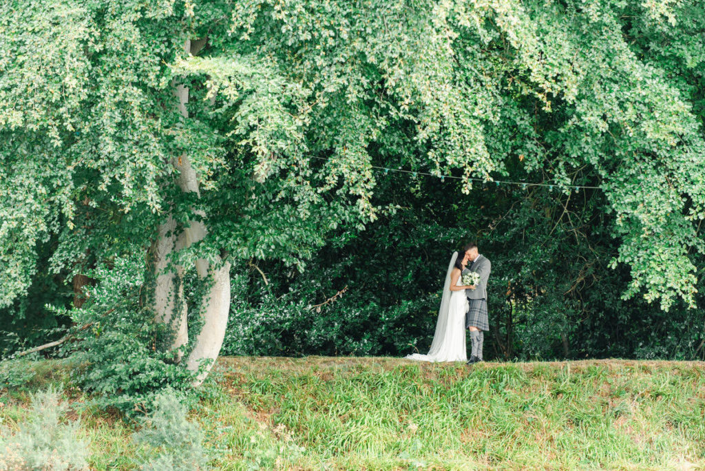 A bride and groom hugging each other standing on a grass bank next to a mature tree with branches overhead