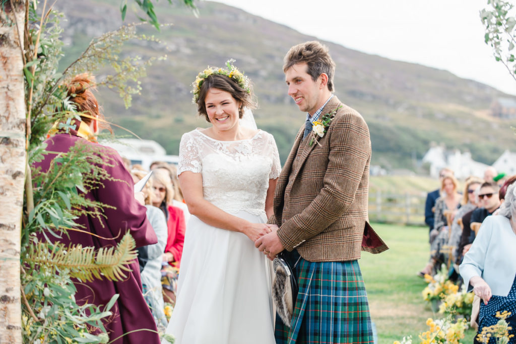 A bride in a white dress and a groom in a jacket and kilt holding hands outdoors with wedding guests seated in the background