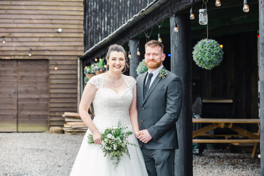 A bride in a white dress holding hands with a groom in a grey suit in front of black and brown farm buildings