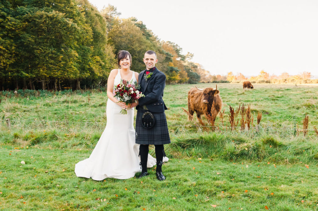 A bride and groom smiling and standing on grass in front of a field with two Highland cows and trees