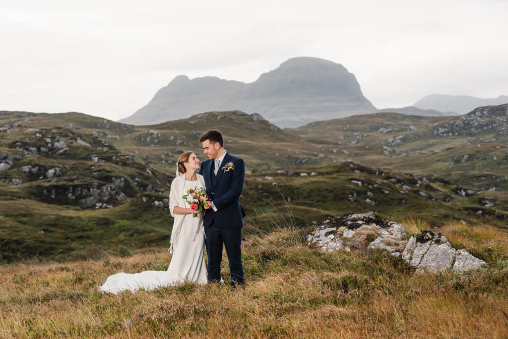 A bride and groom holding flowers and smiling at each other in long grass next to a rock with a mountain in the background