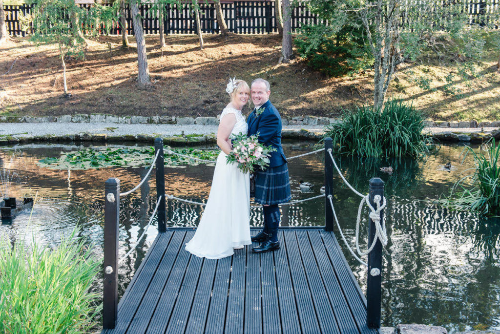A Caucasian bride and groom dressed in a dress and kilt standing on a black jetty above a pond with a path and trees behind