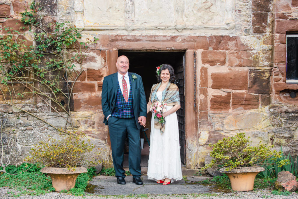 A bride in a white dress and a groom in a trouser suit, holding hands and standing outside the entrance to a sandstone castle