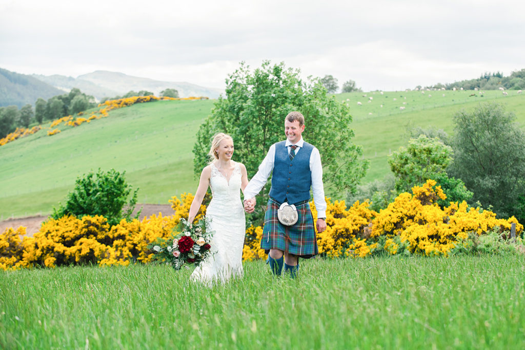 A bride in a white dress holding hands with a groom in a kilt, laughing and walking through grass in front of yellow gorse