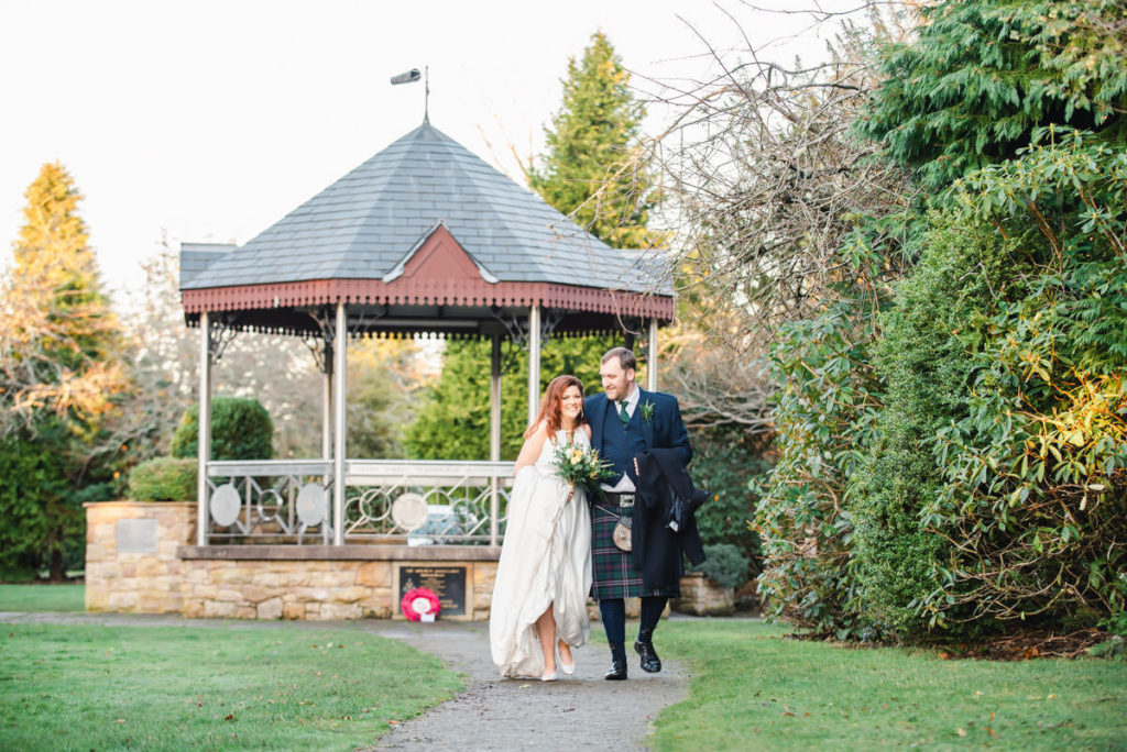 A Caucasian bride and groom smiling and walking on a path next to grass and shrubs with a bandstand in the background