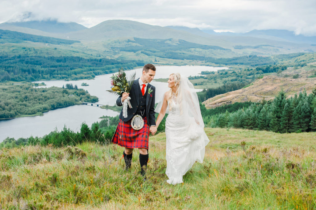 A Caucasian bride and groom holding hands and walking through rough grass with a loch, trees and mountains in the background