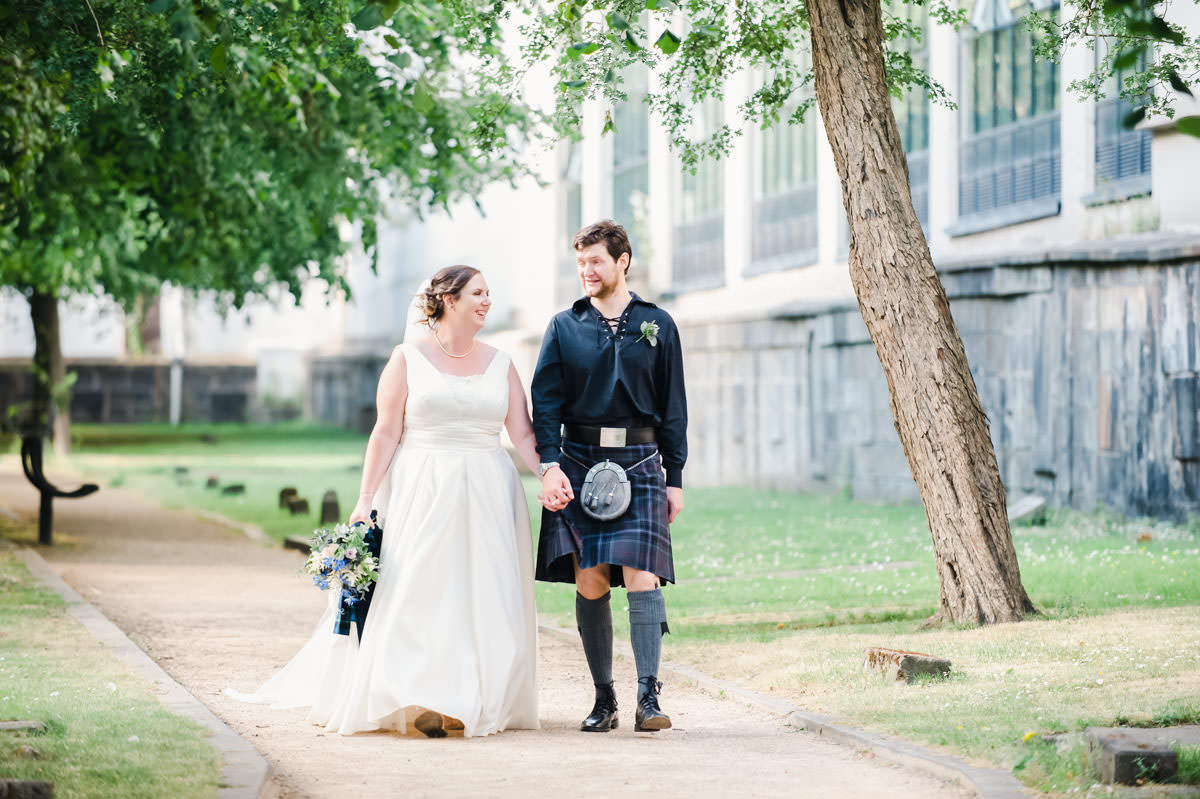 A bride and groom holding hands and walking on a path in urban greenspace with trees, grass and a building in the background