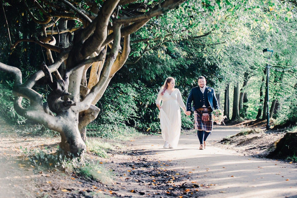 A bride and groom holding hands and walking on a woodland path with sunlight streaming through the trees